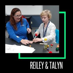 Talyn on the left is wearing a blue, long sleeved shirt, next to Reiley, who is wearing a white long-sleeved shirt. They are building a catapult together and launching pompom balls.