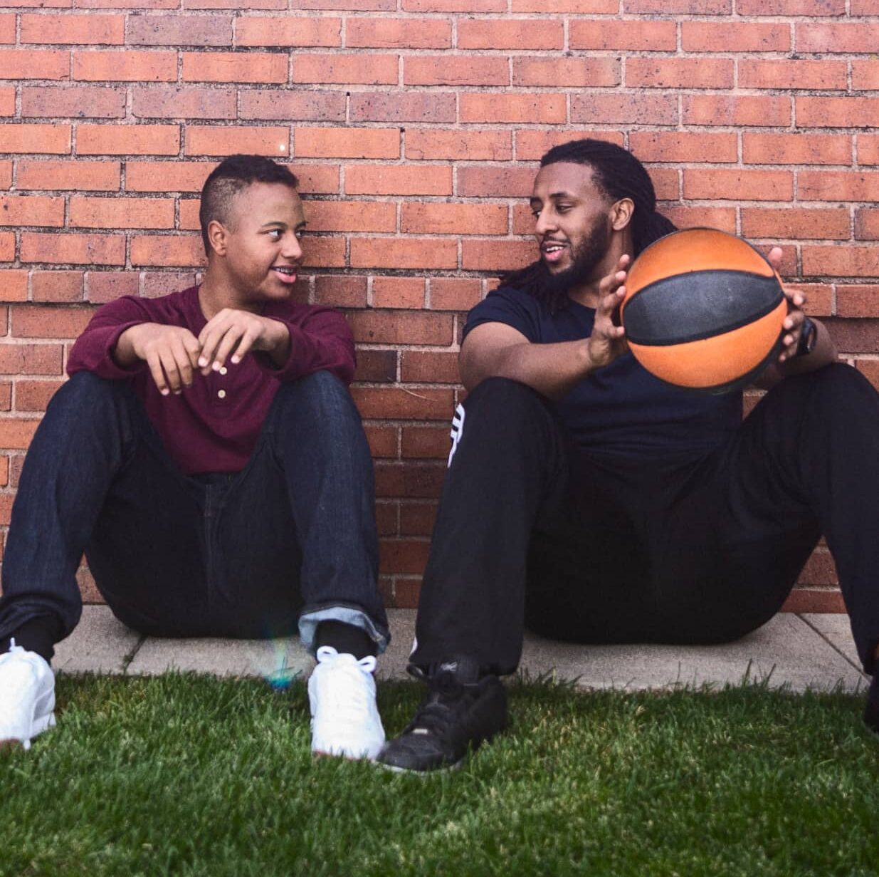 A big and little brother sit outside smiling at each other holding a basketball
