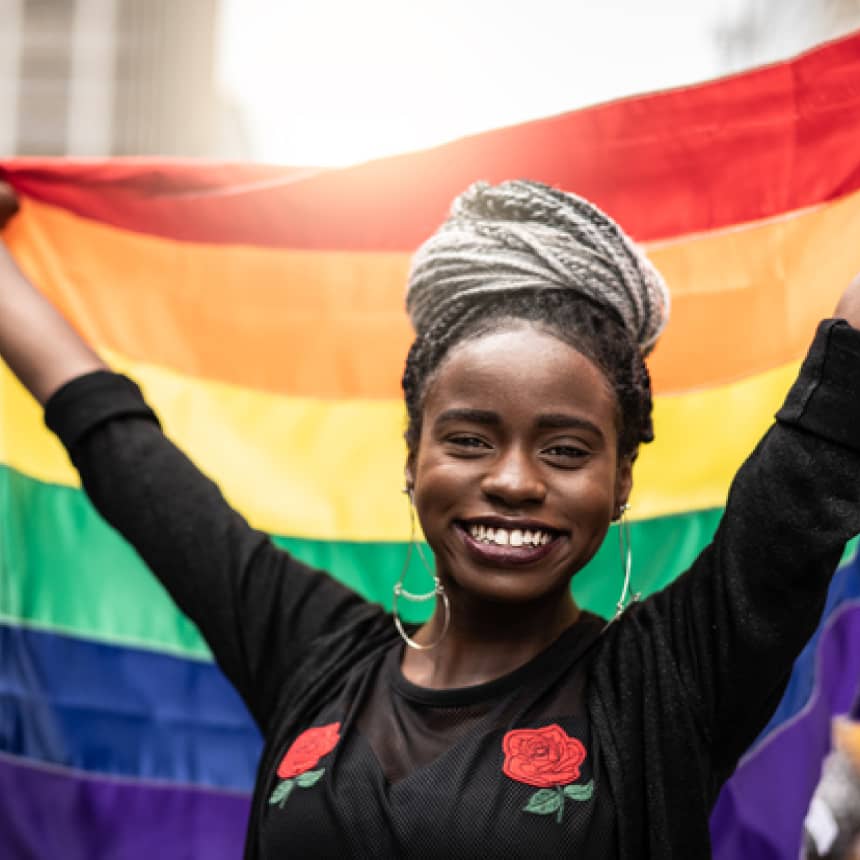 Smiling young black woman holding up a rainbow pride flag behind her.
