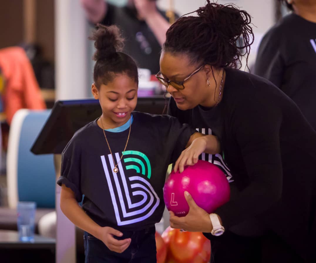 Woman helping a young girl lift her bowling ball at a Bowl for Kids' Sake bowling event.