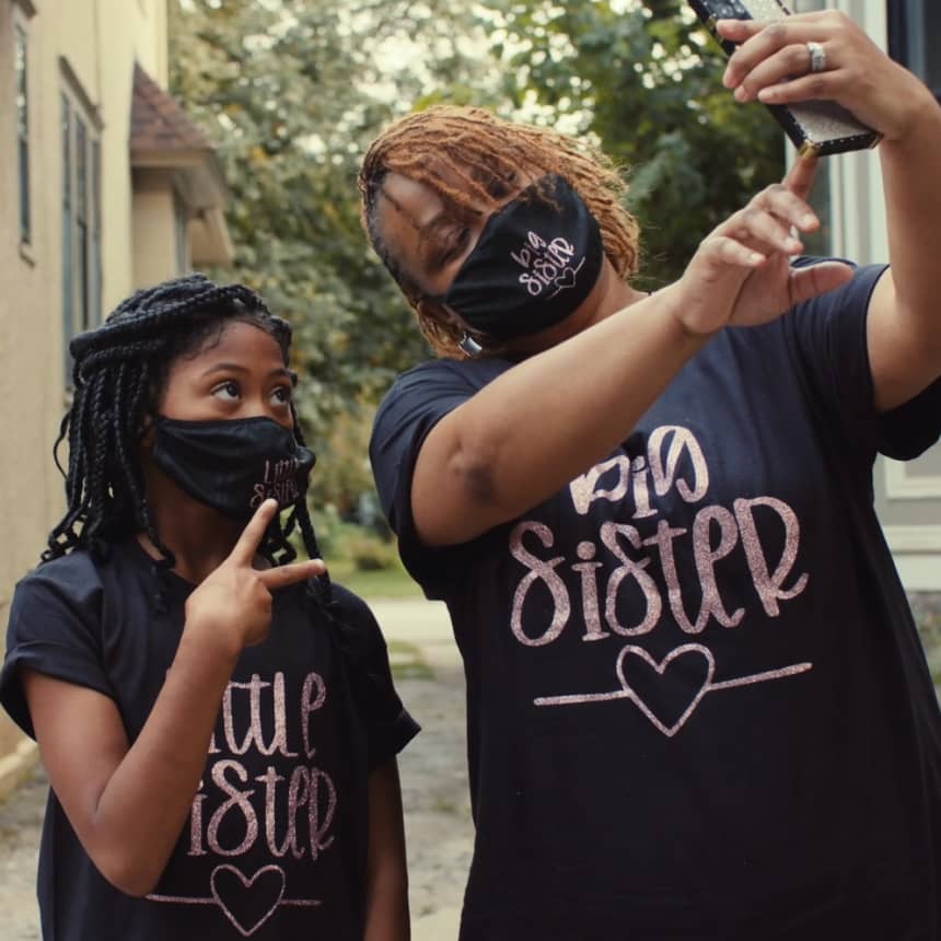 A big sister and little sister wearing matching face masks and t-shirts taking a selfie together.