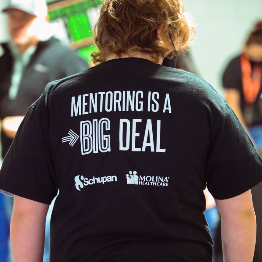 Volunteer with their back to the camera. The back of their shirt features sponsor logos and reads “Mentoring is a big deal.”