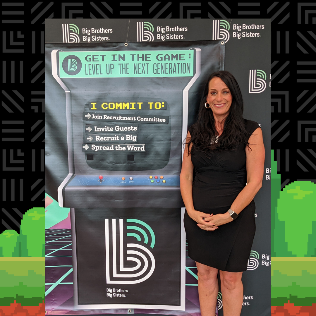 Melissa Johnson stands wearing a black dress, smiling at the camera, standing next to a cutout of a vintage video game console stating the commitments she is making to help find more mentors.