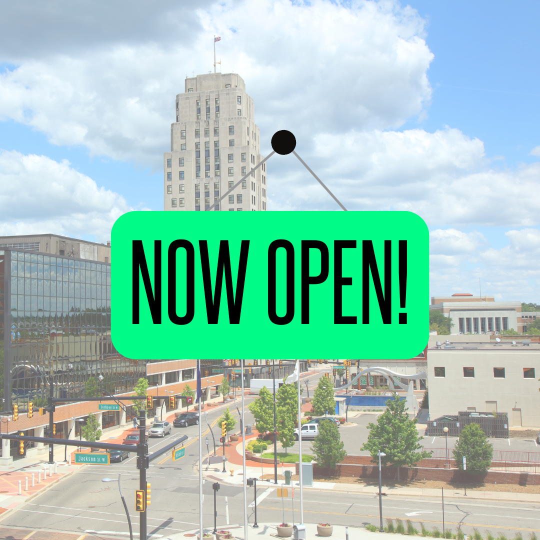 In the background is a photo of downtown Battle Creek. In the foreground is a sign in signature BBBS green that says "Now Open!"