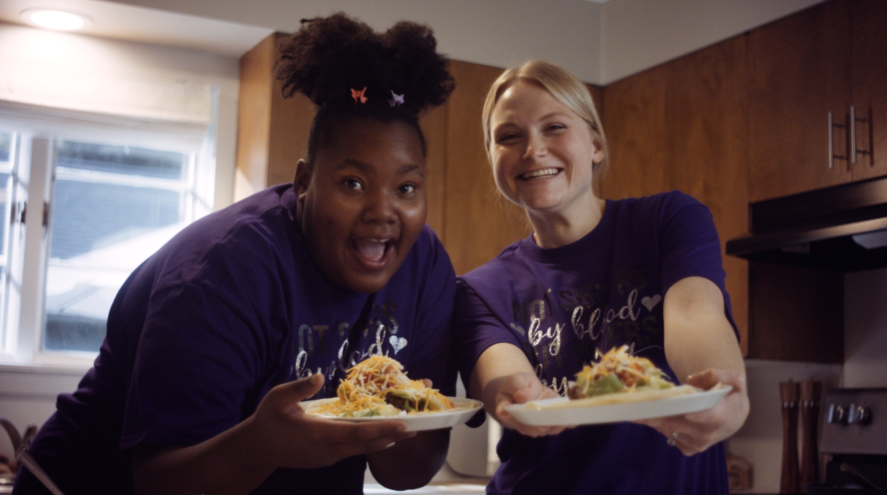 Clare and Jaliyah smile at the camera, holding out plates of tacos.