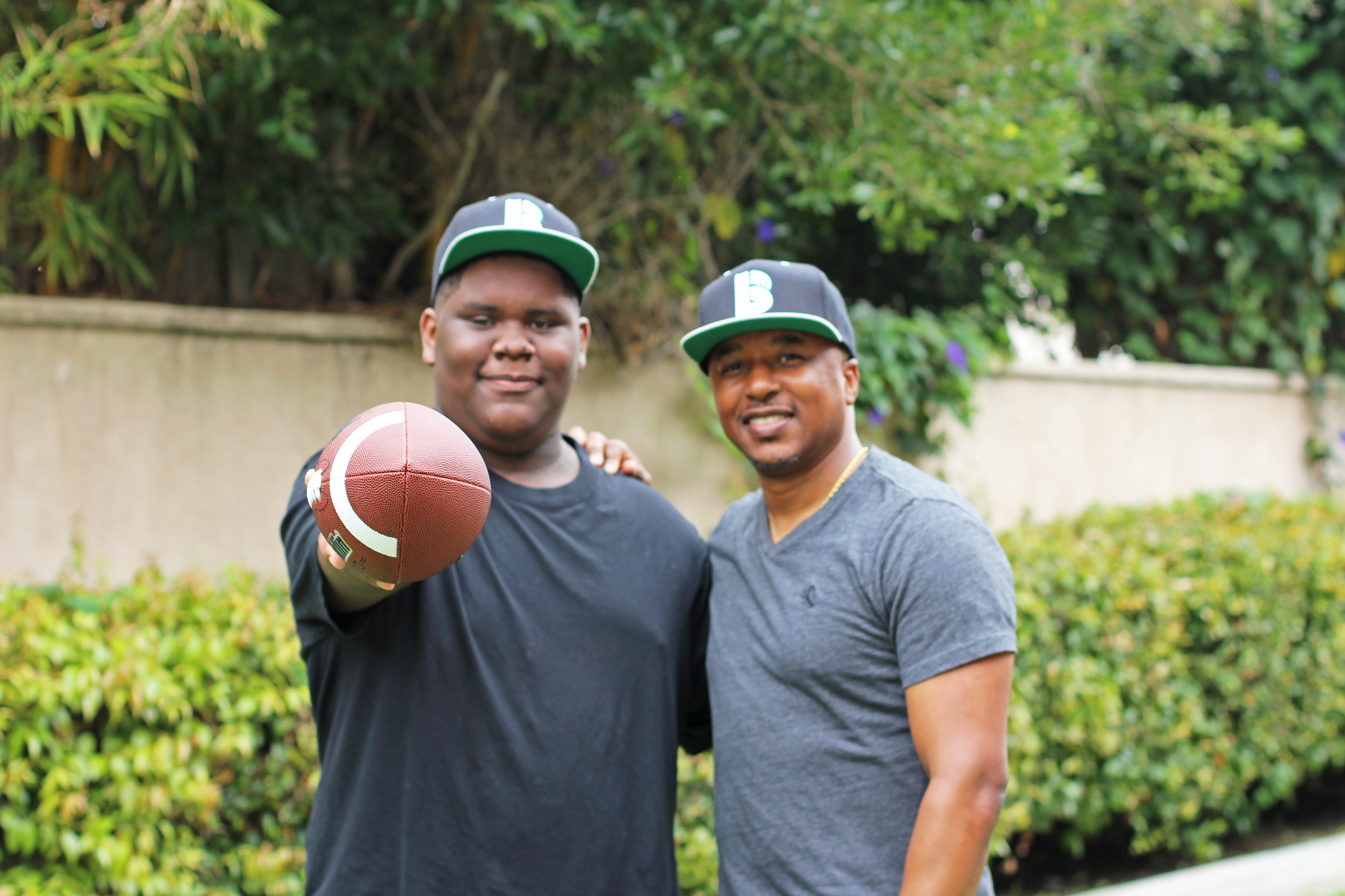 Two people stand next to each other outside, wearing BBBS hats. One is holding a football, outstretched toward the camera.