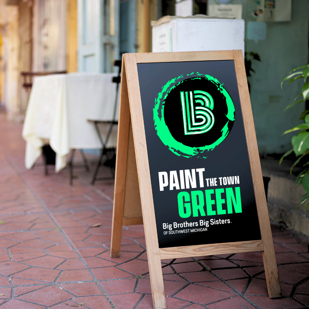 picture of a storefront sidewalk with a chalkboard displaying the Paint the Town Green logo