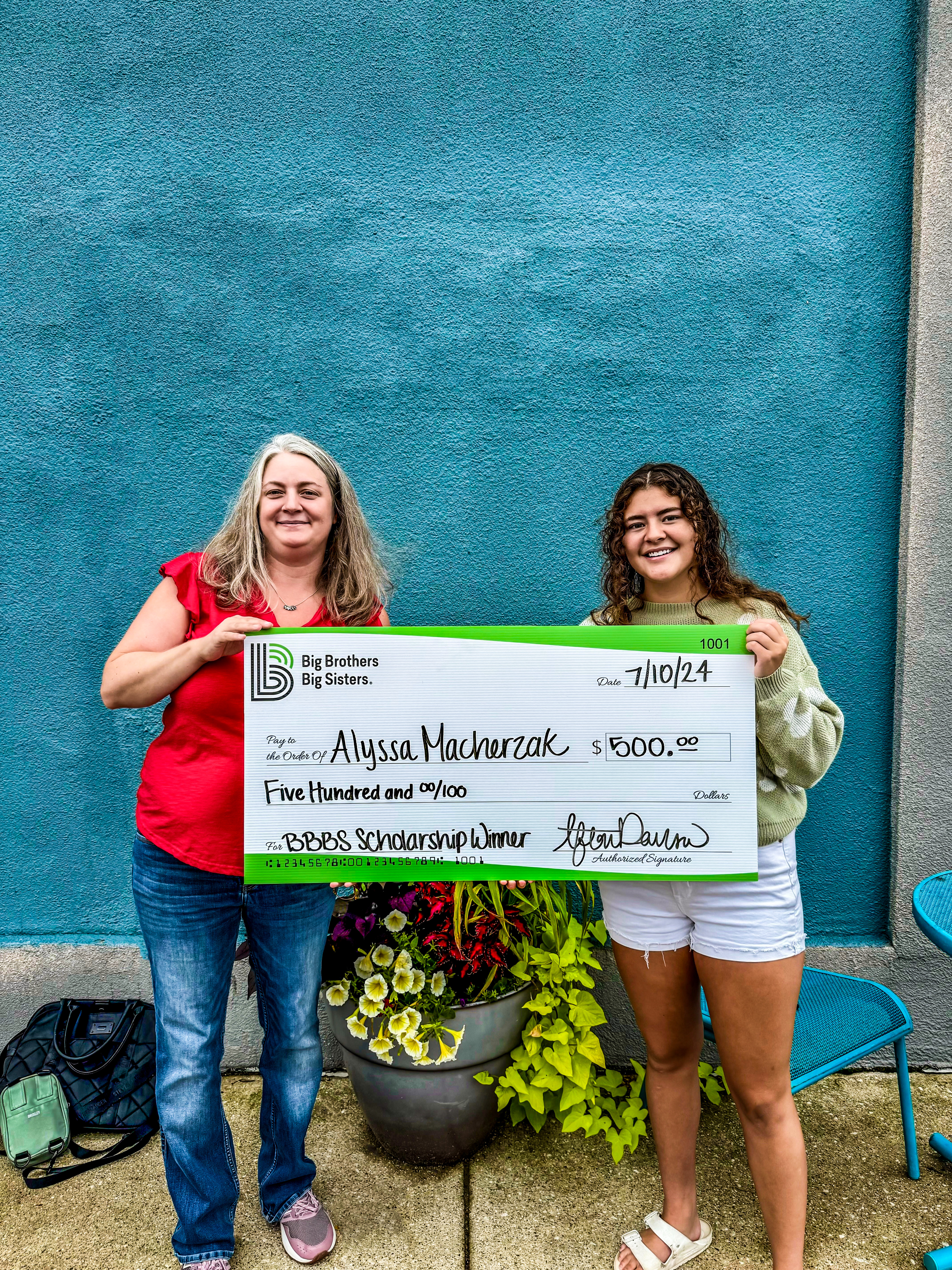 Alyssa and her Big Sister Cydney holding Alyssa's giant check and smiling at the camera in front of a blue wall outside