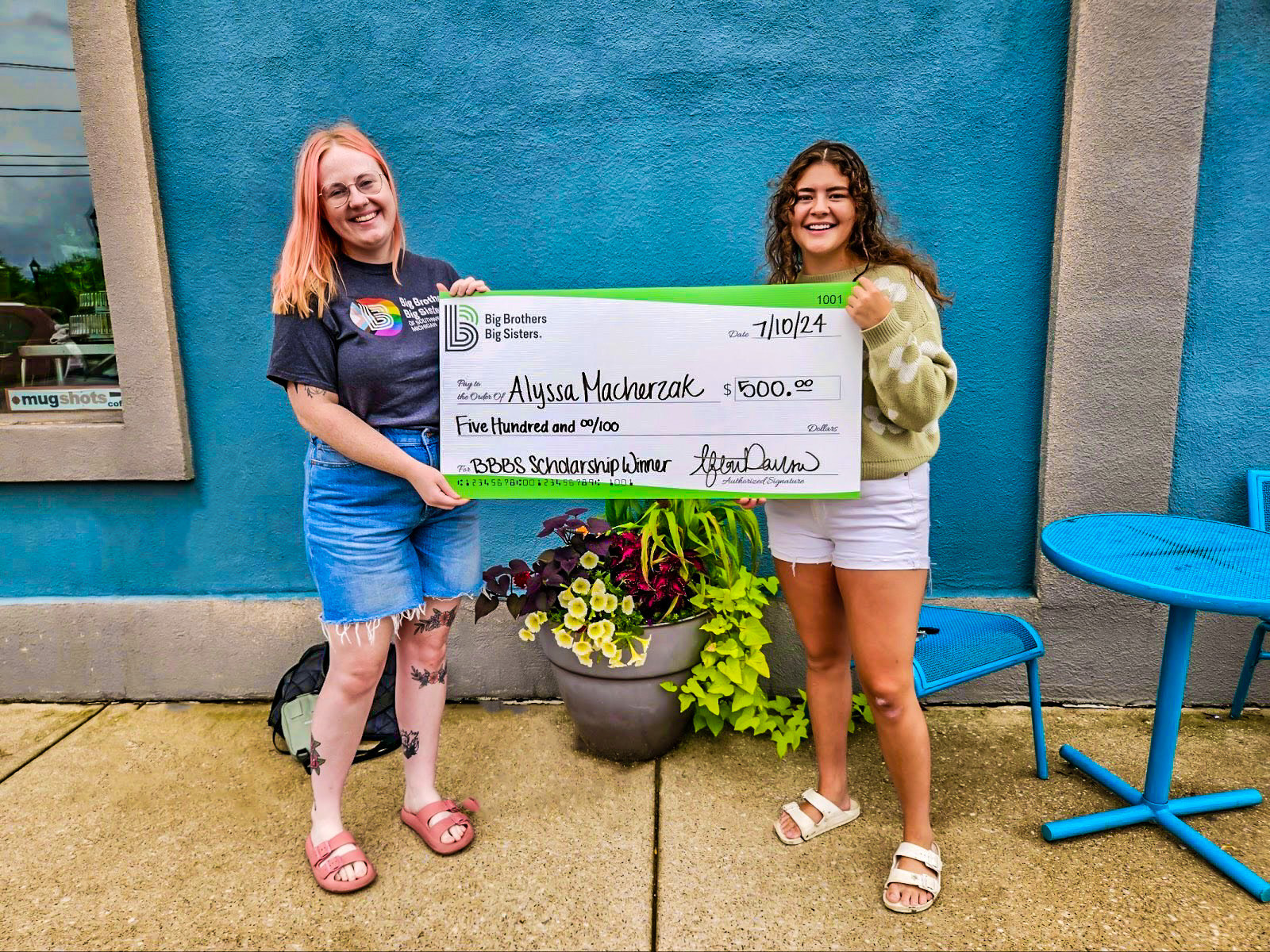 Little Alyssa pictured here with her MRS. They are standing in front of a blue wall, smiling at the camera and holding a giant check made out to her for $500.